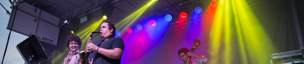 Three men on stage playing and surrounded by a variety of instruments under multicolored lighting.