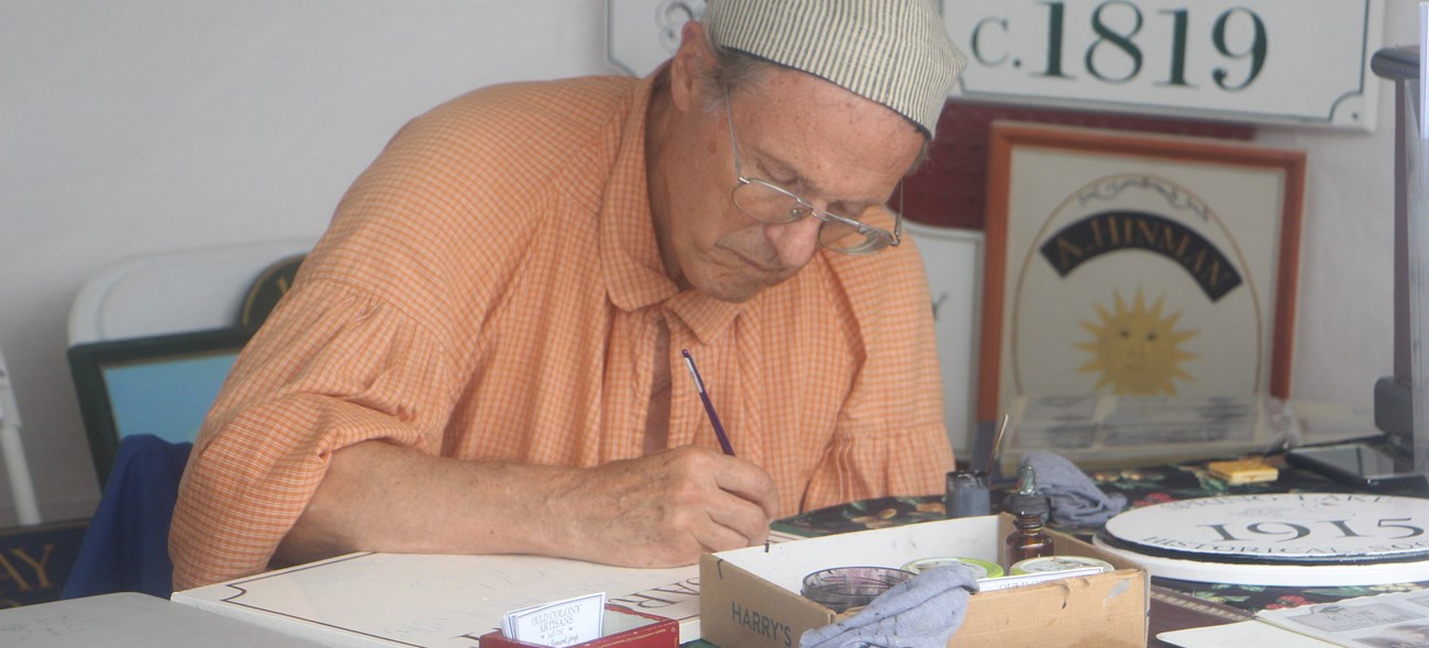 Artist in period clothing paints a sign.