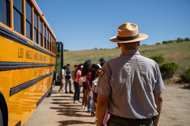 A Park Ranger stands outdoors while students form a line as they get off a school bus.