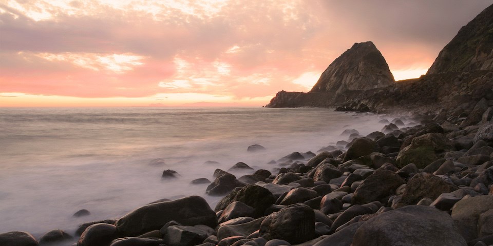 Waves crash upon smooth rocks in the fading evening light, a large rock formation in the distance