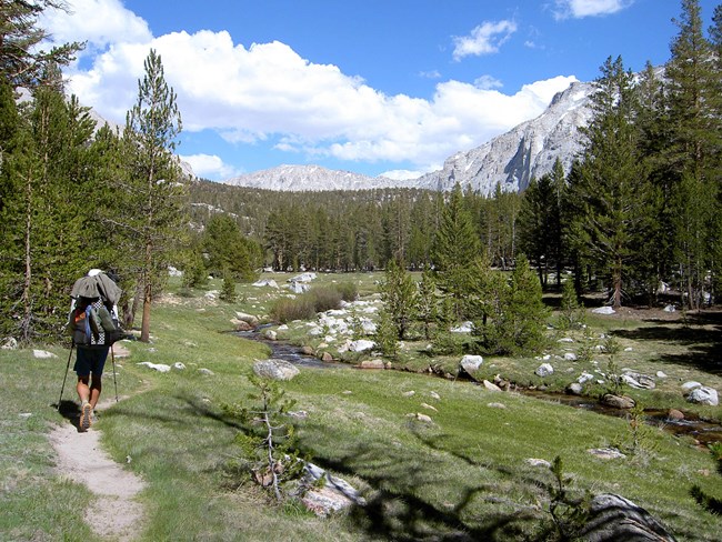 A backpacker hikes along a stream overlooking large mountain vistas.