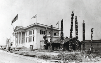 Black and white photograph of the Alaska exhibit at the 1904 Louisiana Purchase Exposition, consisting of a white two story building on the left side of the photo, and a wooden clan house surrounded by seven totem poles on the right.