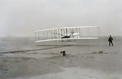 An experimental aircraft launches off a beach with a man standing next to it