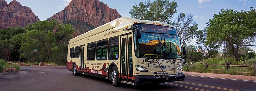 Zion Shuttle Bus A large, new bus in the foreground with sandstone peaks in the background