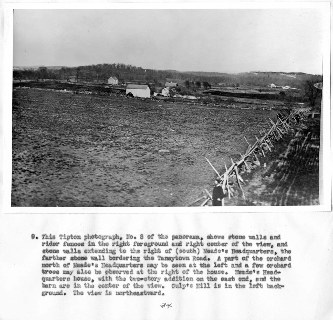 Historic photograph shows a rider fence and stone walls along the edge of a field, with structures and trees in the background.