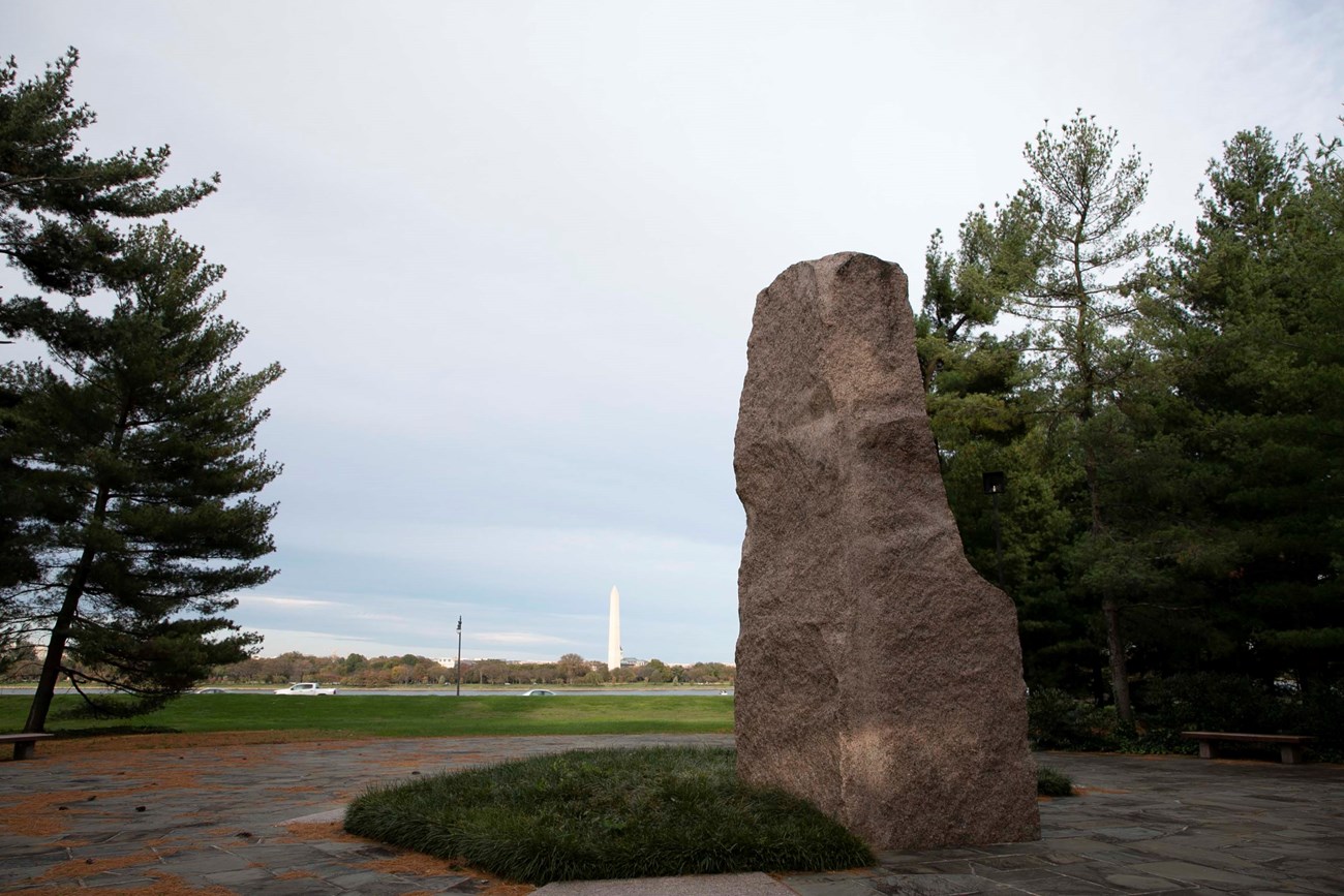 Stone sculpture surrounded by pines and gray flagstones, with the Washington Monument in the rear distance.