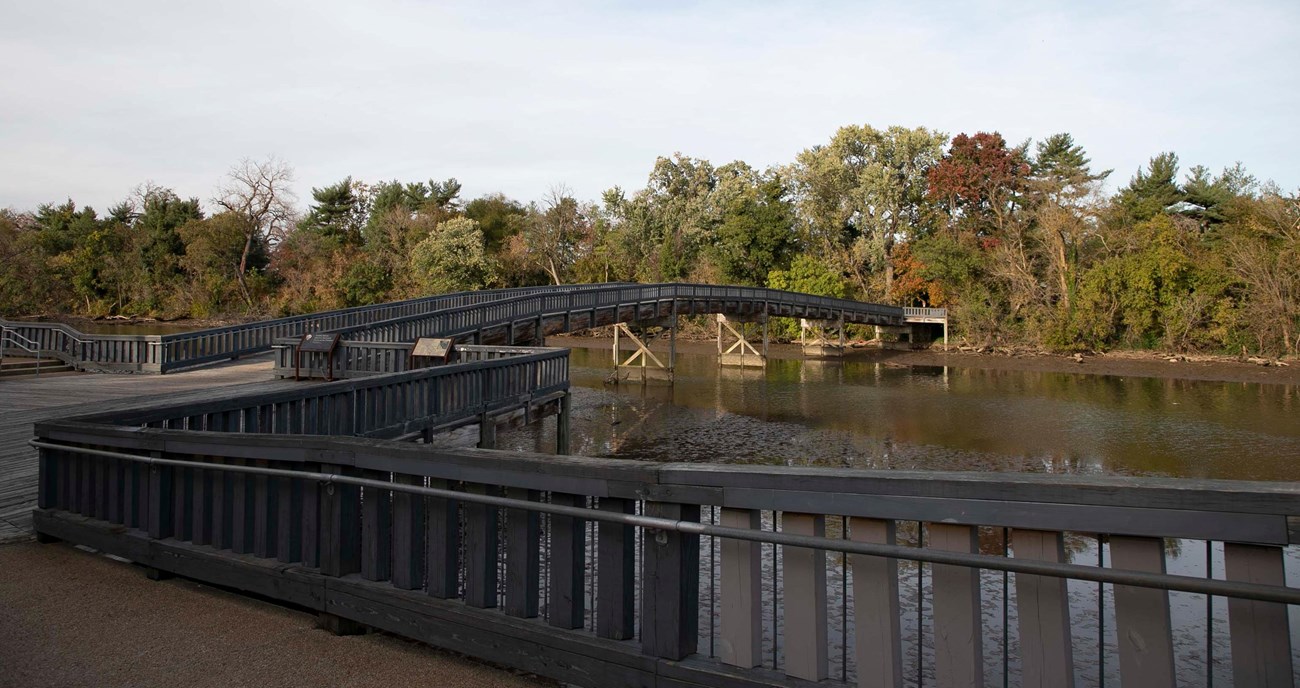 A wooden pedestrian bridge arches slightly to cross a waterway towards a tree-lined shore.