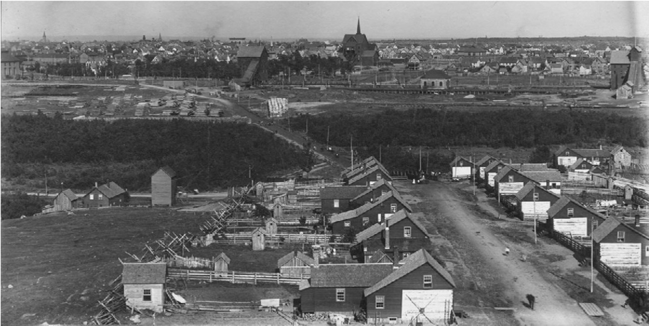 Bridge Street in Swedetown and Calumet beyond,1903 Rows of uniform houses line a street, each with a fenced yard and outbuidlings. Industrial mining structures stand in the distance, near other residential and commercial structures.