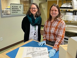 Two women standing side by side examining a file folder of documents. One has blue gloves on.