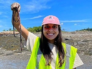 Woman with pink ball cap and green safety vest holds up a large, long worm while she's standing on a pebbly beach