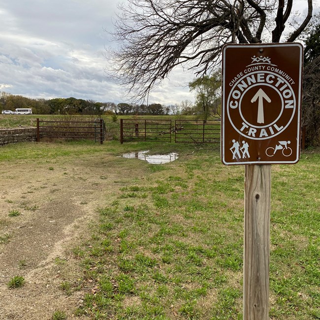 Chase County Community Connection Trail. Grass in foreground with "Connection Trail" sign. Gate leads to more field and a parking area.