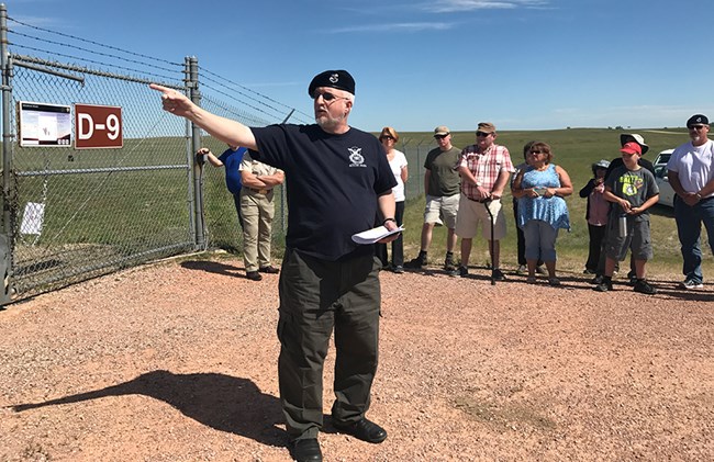 Man gestures to a group standing outside a fenced compound