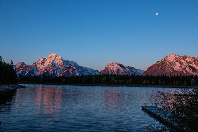 A dock extends into a lake as sunlight hits mountains.