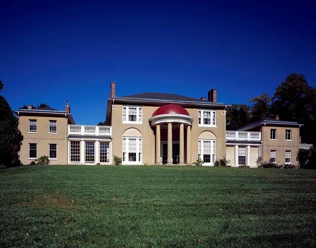 Exterior of Tudor Place. tan walls with red domed porch. Green lawn in the foreground.