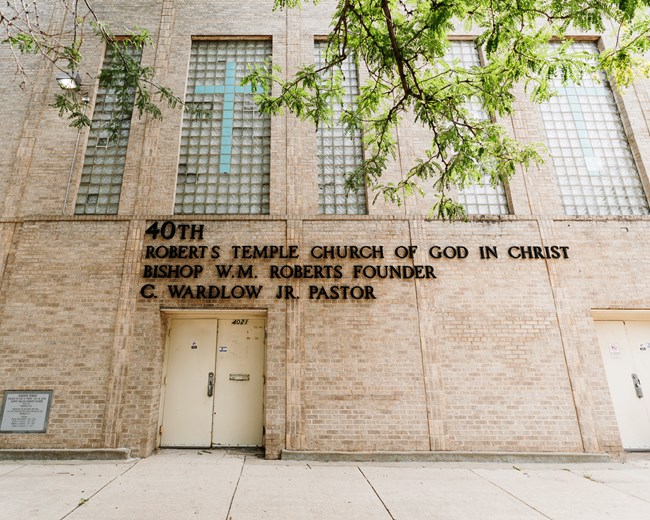 View of a multi-story brick church from the bottom looking toward the top. The name of the church, founder, and current pastor is on the front.