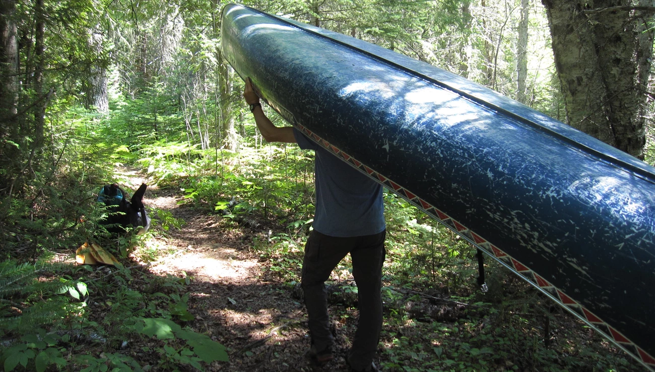 Person Portaging Canoe at Isle Royale A person portages a canoe down a trail surrounded by forest towards a backpack and paddles on the ground.