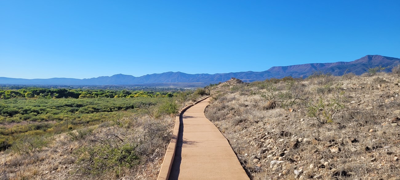 A paved sidewalk along a ridge in the desert. A pueblo is visible on a hilltop in the distance.