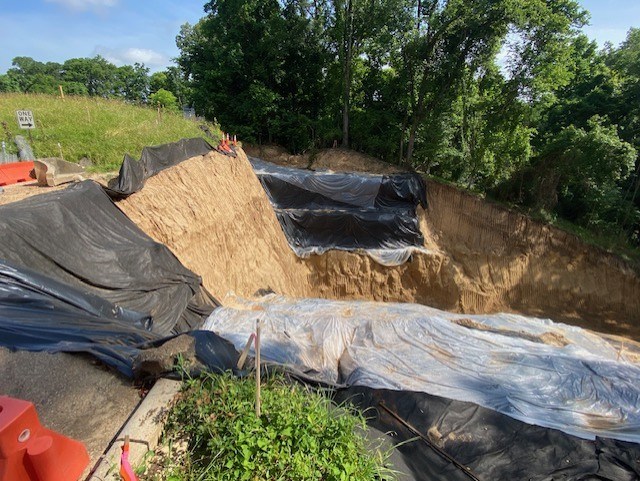 Construction site. Left to right:  High vertical dirt wall at edge of a road with excavated area below partially covered in clear and black tarps