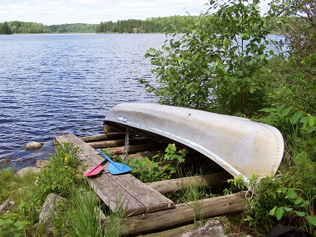 A canoe sits upside down on a wooden rack on the shores of a scenic lake, secured to the rack by a chain. Two paddles lay crossed next to the canoe.