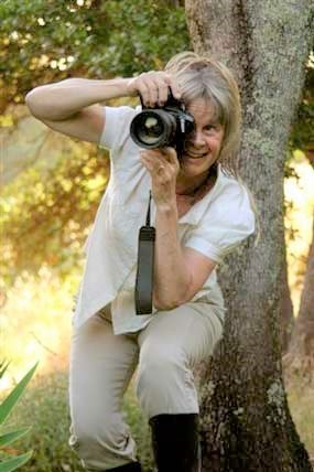 A female artist in residence in white suite pointing a black camera at you, with her face pointing towards you behind the camera.