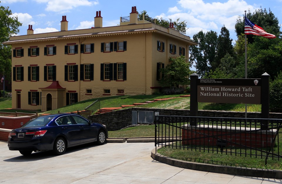 A blue car parked in a parking lot with a large two-story yellow building in the background