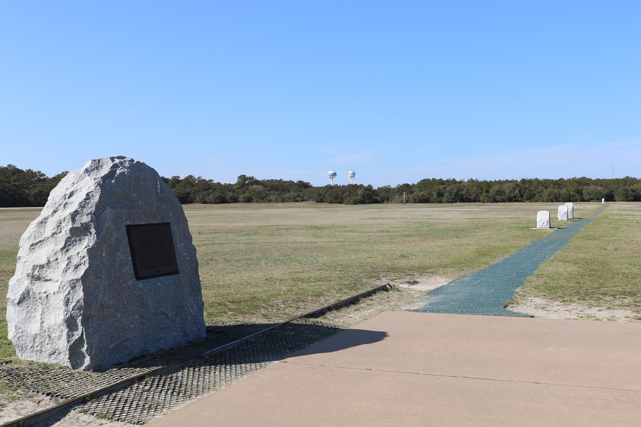 A large boulder sits in a field with smaller, numbered stones in the background. Indicating where the first flights took off and landed.