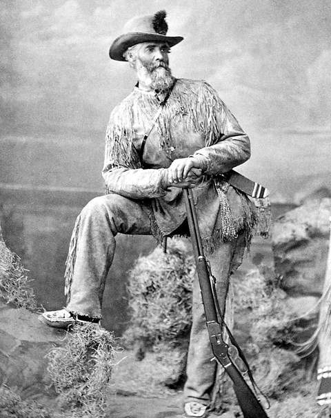 A historic photograph of a man posing in a leather suit with fringes, a hat, and gun