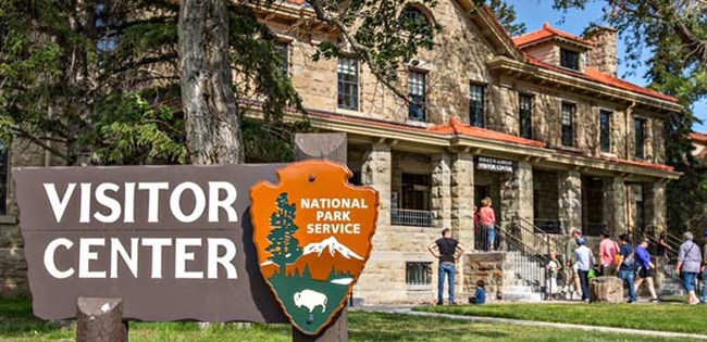 Large brown sign reads "Visitor Center" with NPS arrowhead on it standing before the stone building that is home to the Albright Visitor Center.