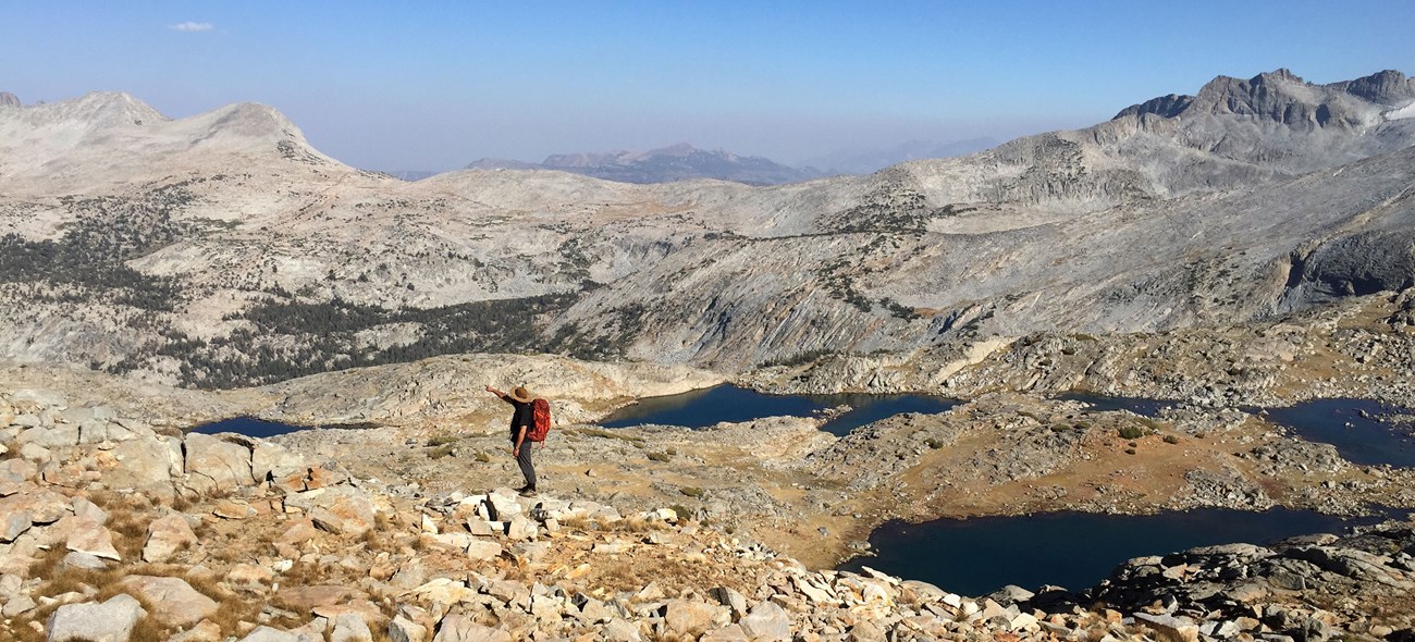 A man stands on a rocky landscape with several small lakes nearby and peaks in the distance