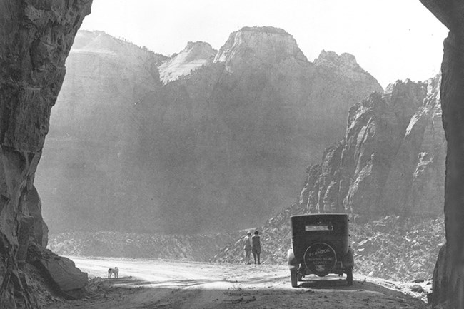 Two people stand near a parked car at the mouth of a tunnel overlooking a deep canyon