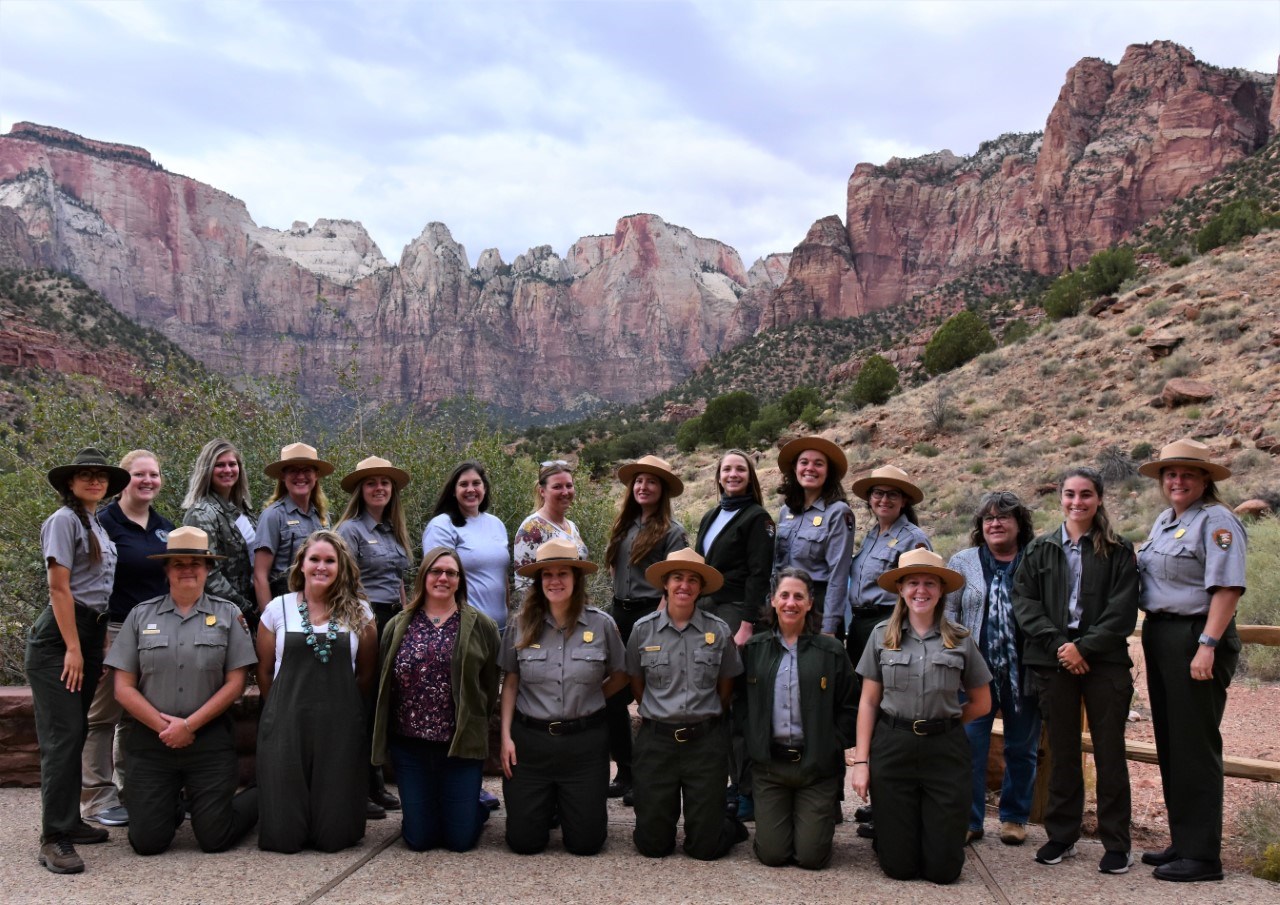 21 women in uniform and formal clothing standing and kneeling in two rows smiling at the camera with tall red sandstone towers in the background